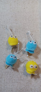 Four small decorative paper fasteners shaped like blue and yellow Creatures with black and white googly eyes, arranged on a beige textured fabric background.