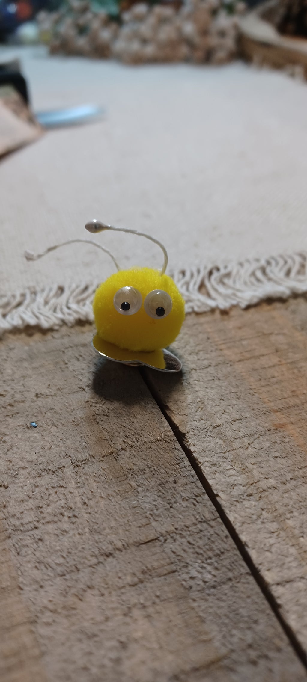 A small yellow novelty decorative paper fasteners shaped like blue and yellow Creatures with black and white googly eyes and thin metal antennae, resting on a wooden surface.