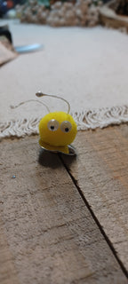 A small yellow novelty decorative paper fasteners shaped like blue and yellow Creatures with black and white googly eyes and thin metal antennae, resting on a wooden surface.