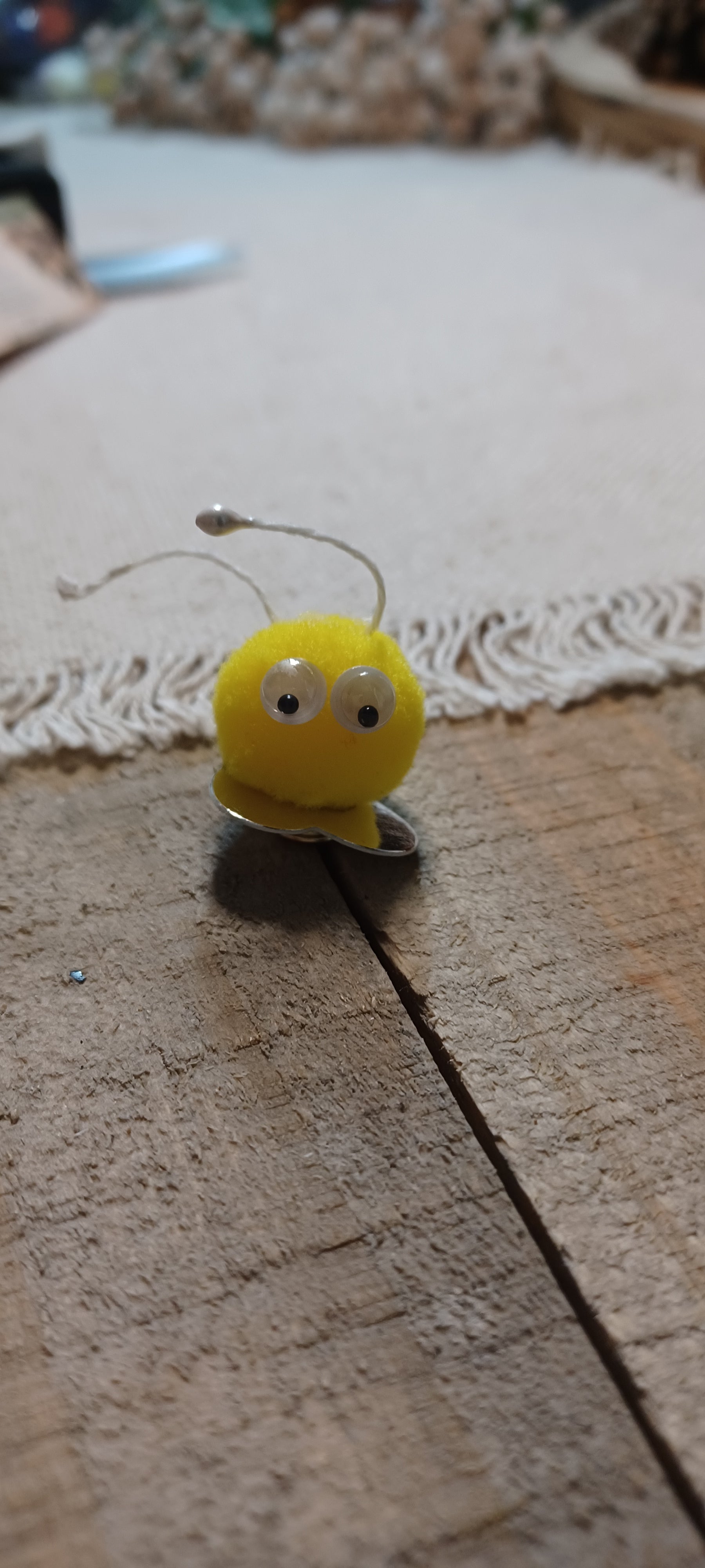 A small yellow novelty decorative paper fasteners shaped like blue and yellow Creatures with black and white googly eyes and thin metal antennae, resting on a wooden surface.