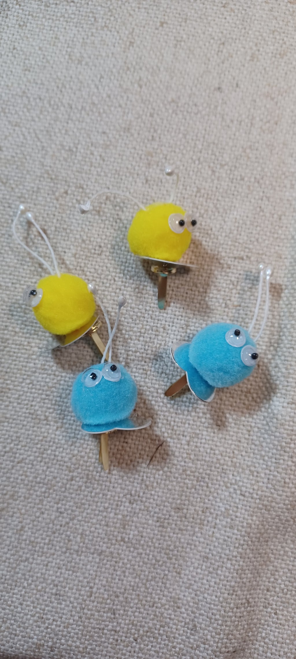 Four small decorative paper fasteners shaped like blue and yellow Creatures with black and white googly eyes arranged on a beige textured fabric background.