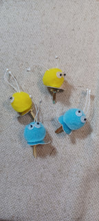Four small decorative paper fasteners shaped like blue and yellow Creatures with black and white googly eyes arranged on a beige textured fabric background.
