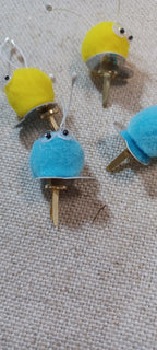 four small decorative paper fasteners shaped like cartoon creatures, with yellow and blue fuzzy bodies, metallic feet, and small black googly eyes, placed on a beige textured fabric background.