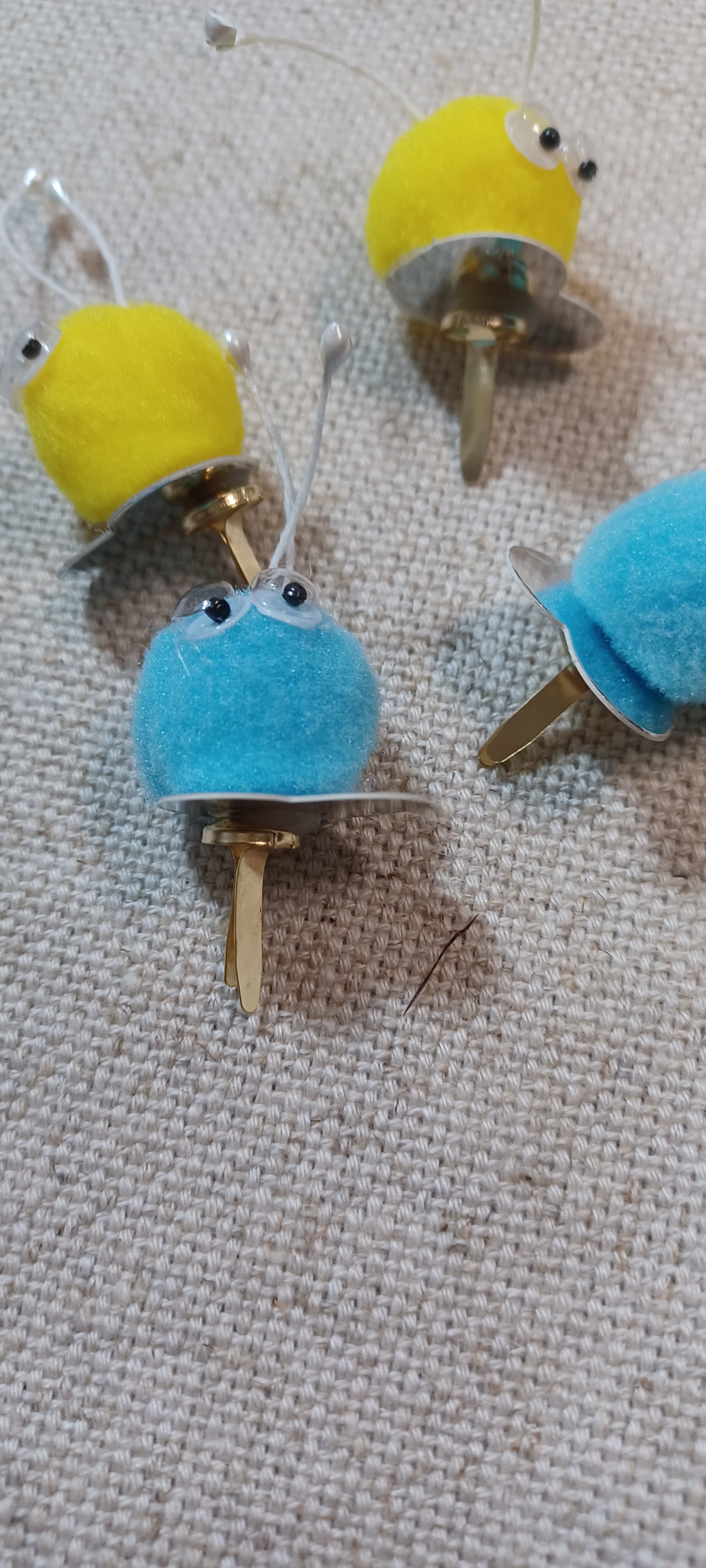 four small decorative paper fasteners shaped like cartoon creatures, with yellow and blue fuzzy bodies, metallic feet, and small black googly eyes, placed on a beige textured fabric background.