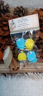 A clear plastic bag containing four decorative paper fasteners shaped like blue and yellow Creatures with black and white googly eyes, labeled 'Butterfly in the Attic' on a kraft header card. The package is styled on a wooden surface with pinecones, cinnamon sticks, and a stone.