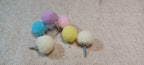 Five small felt pom-poms in pastel colors including blue, purple, pink, yellow, and off-white, each attached to a Brad, arranged on a beige textured fabric background.