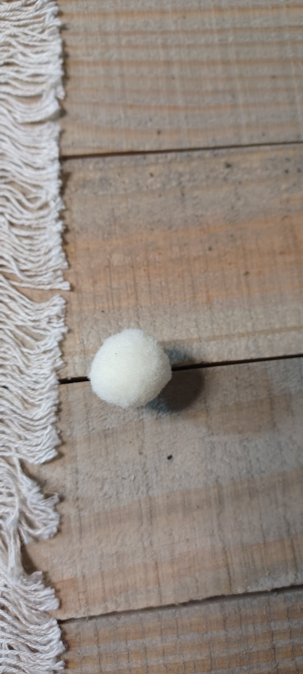 A small white felt pom-pom decorative paper fasteners, placed on a rustic wooden surface next to a cream-colored fringed textile.