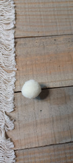 A small white felt pom-pom decorative paper fasteners, placed on a rustic wooden surface next to a cream-colored fringed textile.