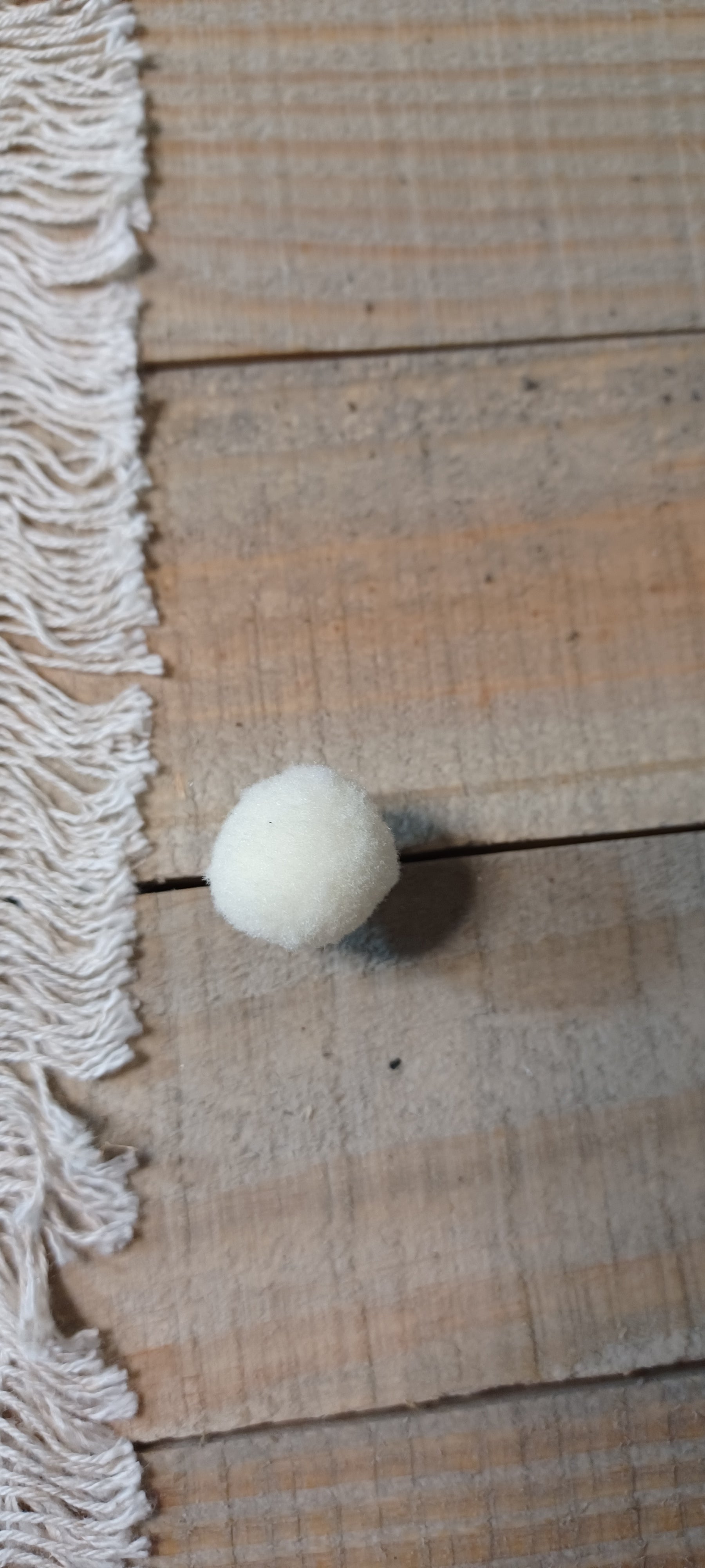 A small white felt pom-pom decorative paper fasteners, placed on a rustic wooden surface next to a cream-colored fringed textile.