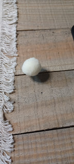 A small white pom-pom ball decorative paper fasteners, resting on a rustic wooden plank surface next to a white fringed textile.