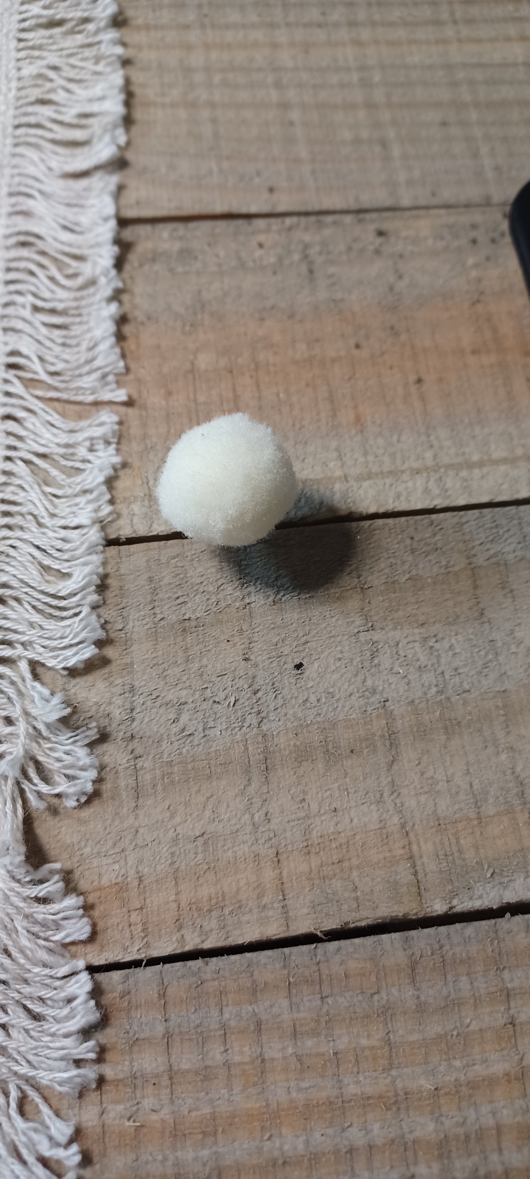 A small white pom-pom ball decorative paper fasteners, resting on a rustic wooden plank surface next to a white fringed textile.