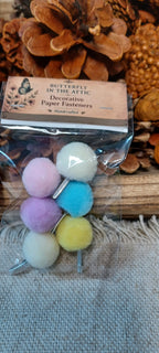A clear plastic bag containing six decorative paper fasteners with fluffy pom-poms in pastel pink, lavender, blue, yellow, and cream. The product is labeled 'Butterfly in the Attic' and is displayed on a rustic wooden surface with pinecones and burlap fabric.
