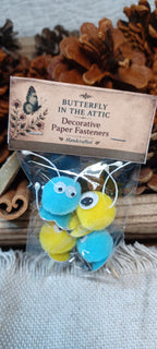 A clear plastic bag containing three decorative paper fasteners shaped like blue and yellow creatures with googly eyes, attached to a printed paper header card that reads 'BUTTERFLY IN THE ATTIC Decorative Paper Fasteners'. The package is photographed on a textured fabric surface with pinecones and wood in the background.