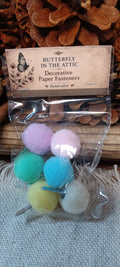 A clear plastic bag containing six colorful decorative paper fasteners, labeled 'BUTTERFLY IN THE ATTIC' on a rustic wood and burlap background.