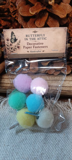 A clear plastic bag containing six colorful decorative paper fasteners, labeled 'BUTTERFLY IN THE ATTIC' on a rustic wood and burlap background.