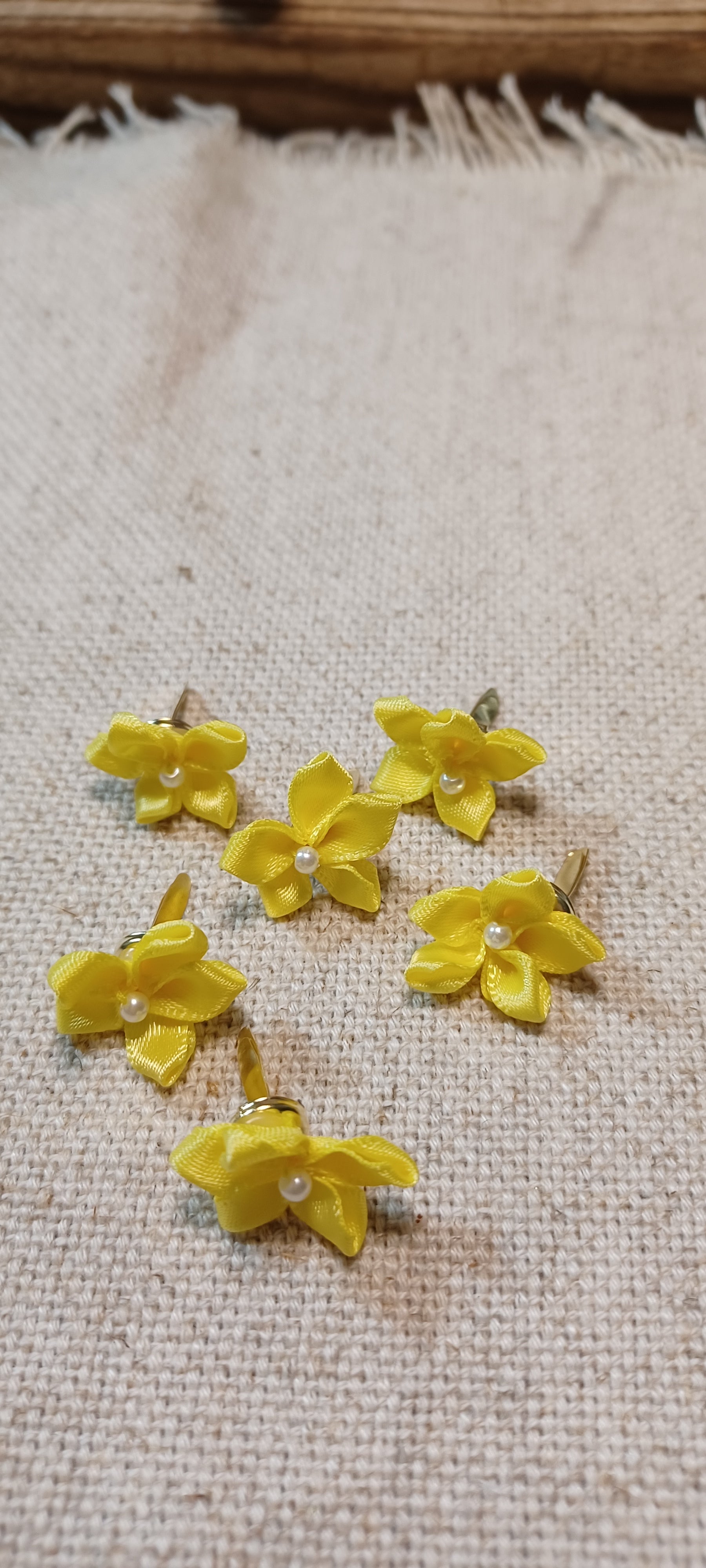 Six yellow daffodil flower paper fasteners with small white pearl centers, arranged on a beige linen fabric background.