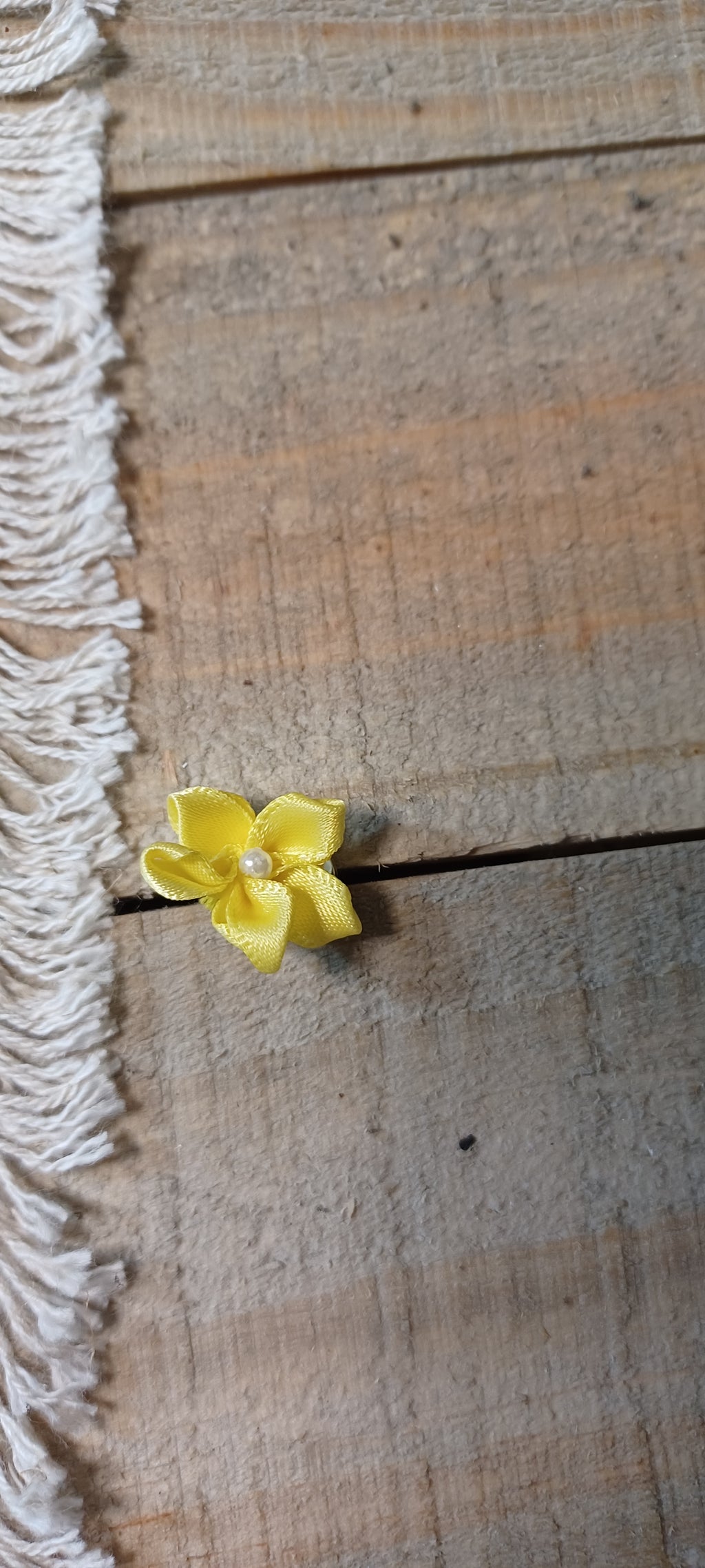A small yellow flower with a pearl center paper fasteners rests on a rustic wooden plank surface next to a white fringed textile.