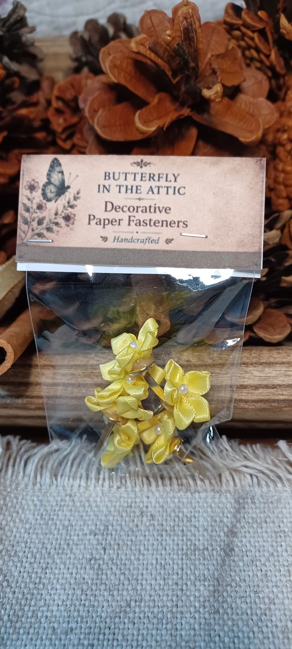 A clear plastic bag containing yellow flower-shaped paper fasteners, with a kraft paper label that reads 'BUTTERFLY IN THE ATTIC Decorative Paper Fasteners'. The product is displayed on a rustic wooden surface with pinecones and a gray textured fabric background.