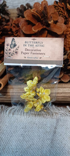 A clear plastic bag containing yellow flower-shaped paper fasteners, with a kraft paper label that reads 'BUTTERFLY IN THE ATTIC Decorative Paper Fasteners'. The product is displayed on a rustic wooden surface with pinecones and a gray textured fabric background.