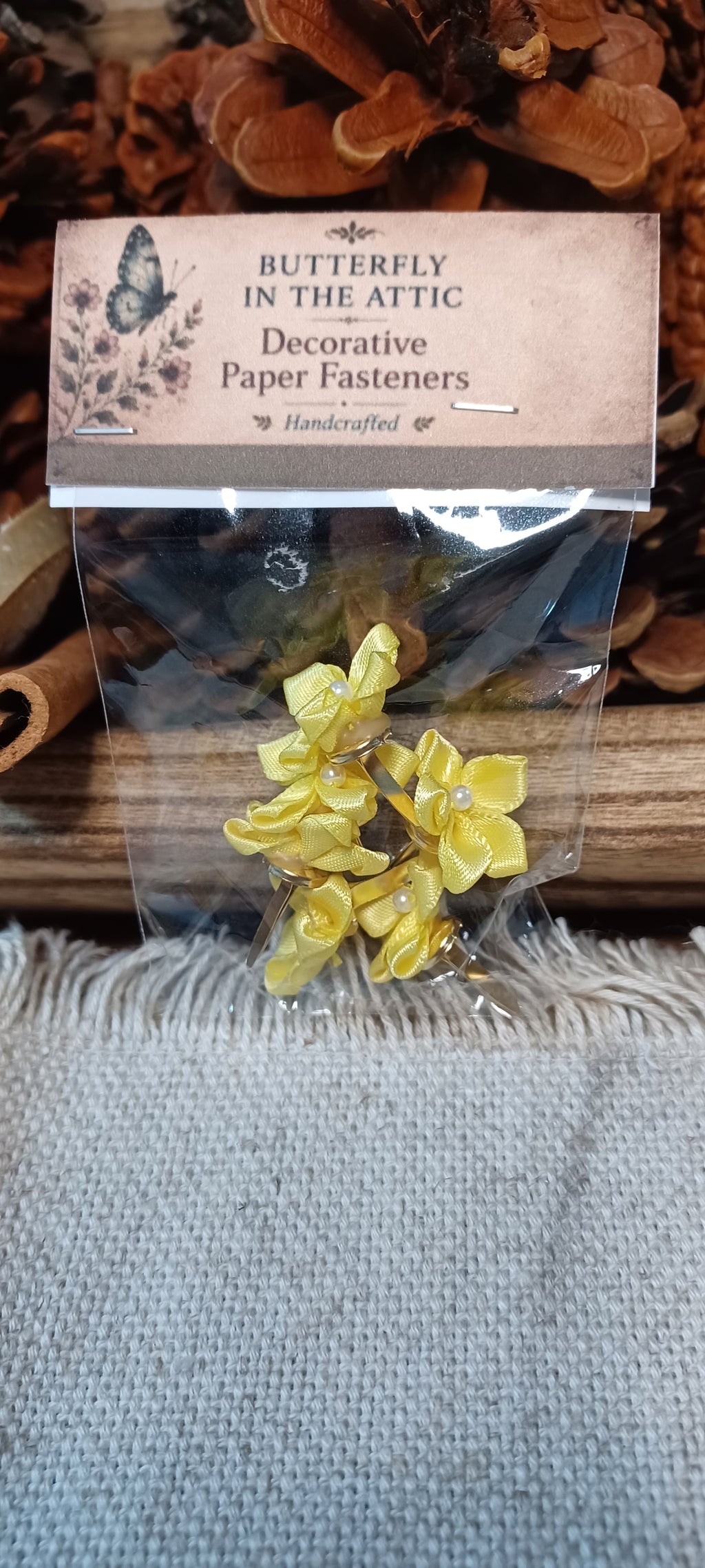 Yellow flower paper fasteners in a clear plastic bag with a kraft paper label, displayed on a rustic wooden surface with dried flowers and a burlap cloth.