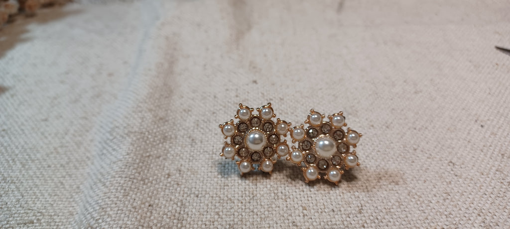 A pair of vintage-style floral decorative paper fasteners with white faux pearls and gold-toned metal, resting on a textured beige linen fabric background.