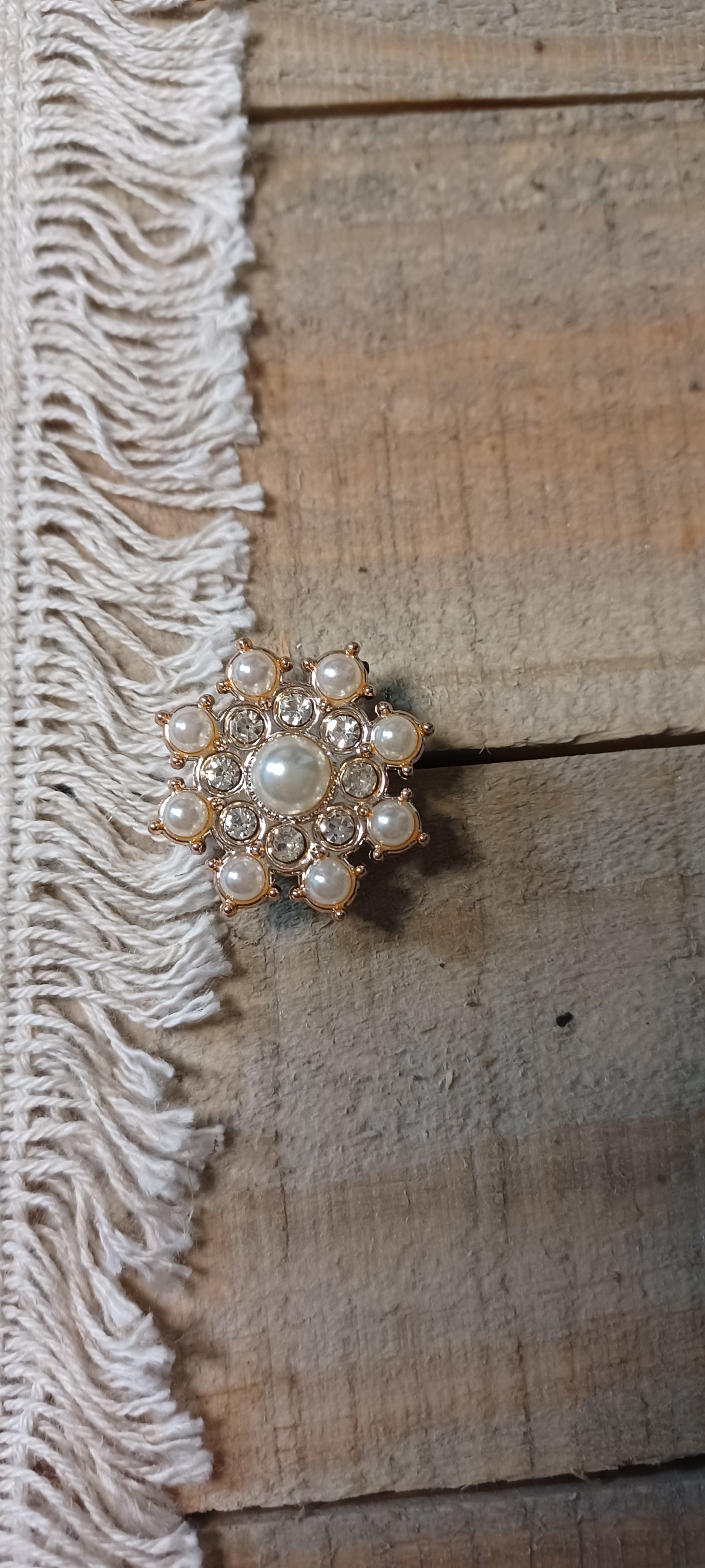 A vintage-style floral paper fastener with a central pearl and surrounding faux pearls and clear rhinestones in a gold-tone setting, displayed on a rustic wooden surface next to a white fringed textile.