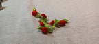 A pair of handmade decorative paper fasteners featuring small red fabric roses with green ribbon leaves, attached to gold-tone wire stems, displayed on a textured beige fabric background.