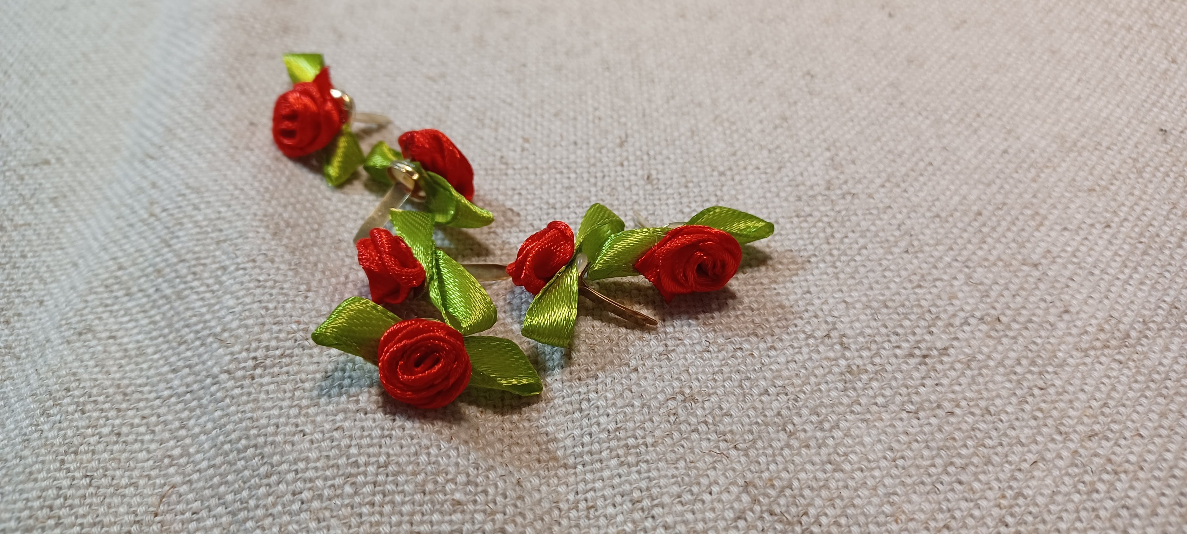 A pair of handmade rose decorative paper fasteners made from rolled red fabric petals with green ribbon leaves, displayed on a textured beige linen background.