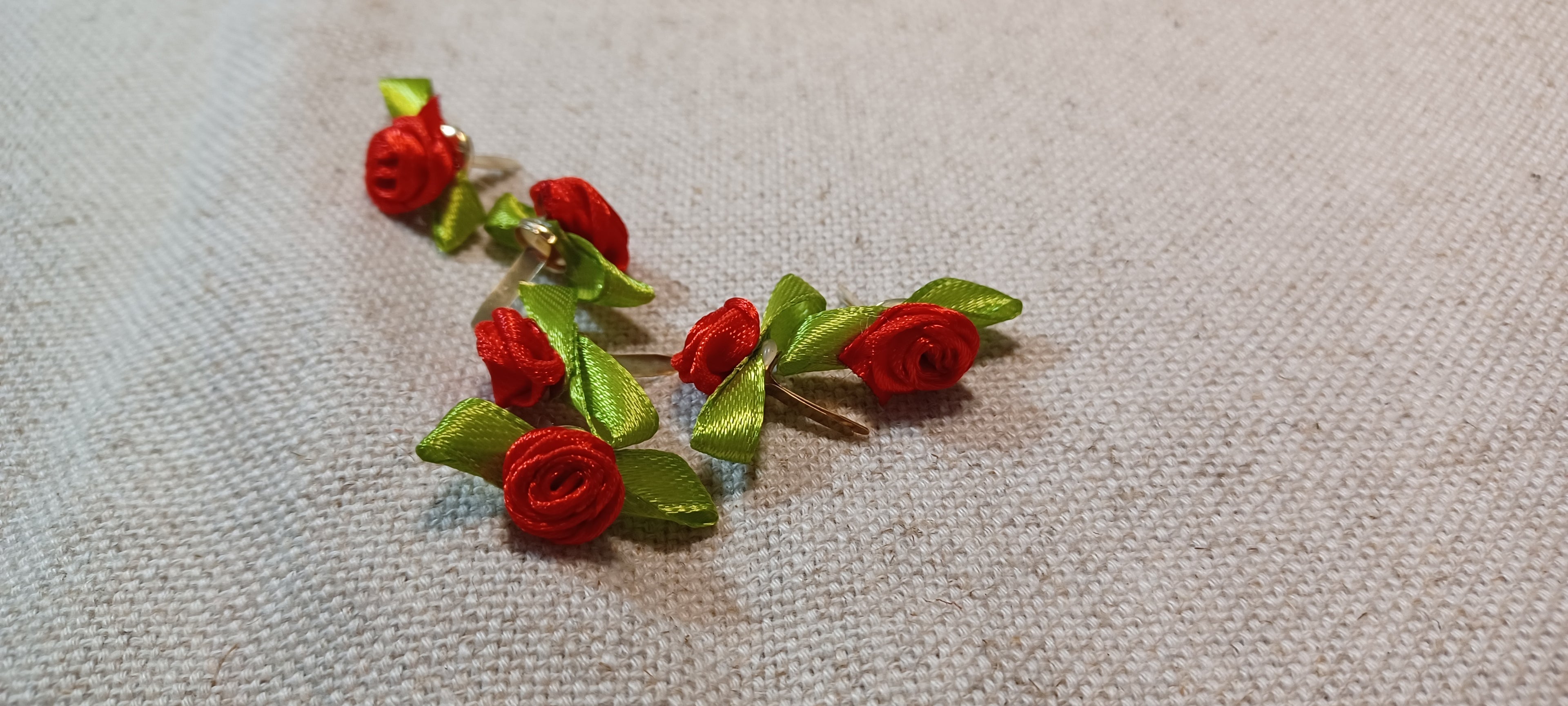 A pair of handmade red ribbon rose decorative paper fasteners with green ribbon leaves, displayed on a textured beige linen fabric background.