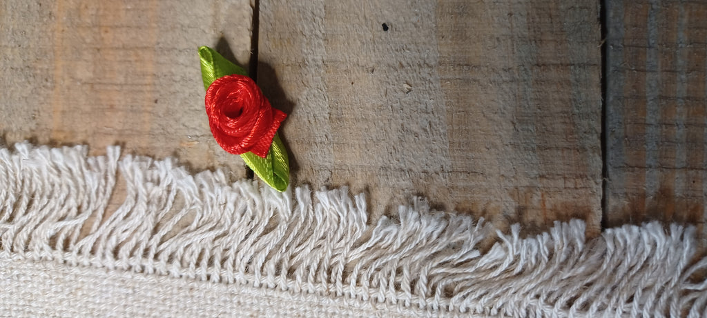 A small embroidered red rose with green leaves decorative paper fasteners on a rustic wooden surface above a white fringed textile.