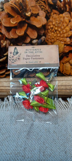 A small clear plastic bag containing red and green decorative paper fasteners, with a paper label reading 'BUTTERFLY IN THE ATTIC' and 'Decorative Paper Fasteners'. The bag is placed on a gray fabric surface with pinecones and a wooden block in the background.