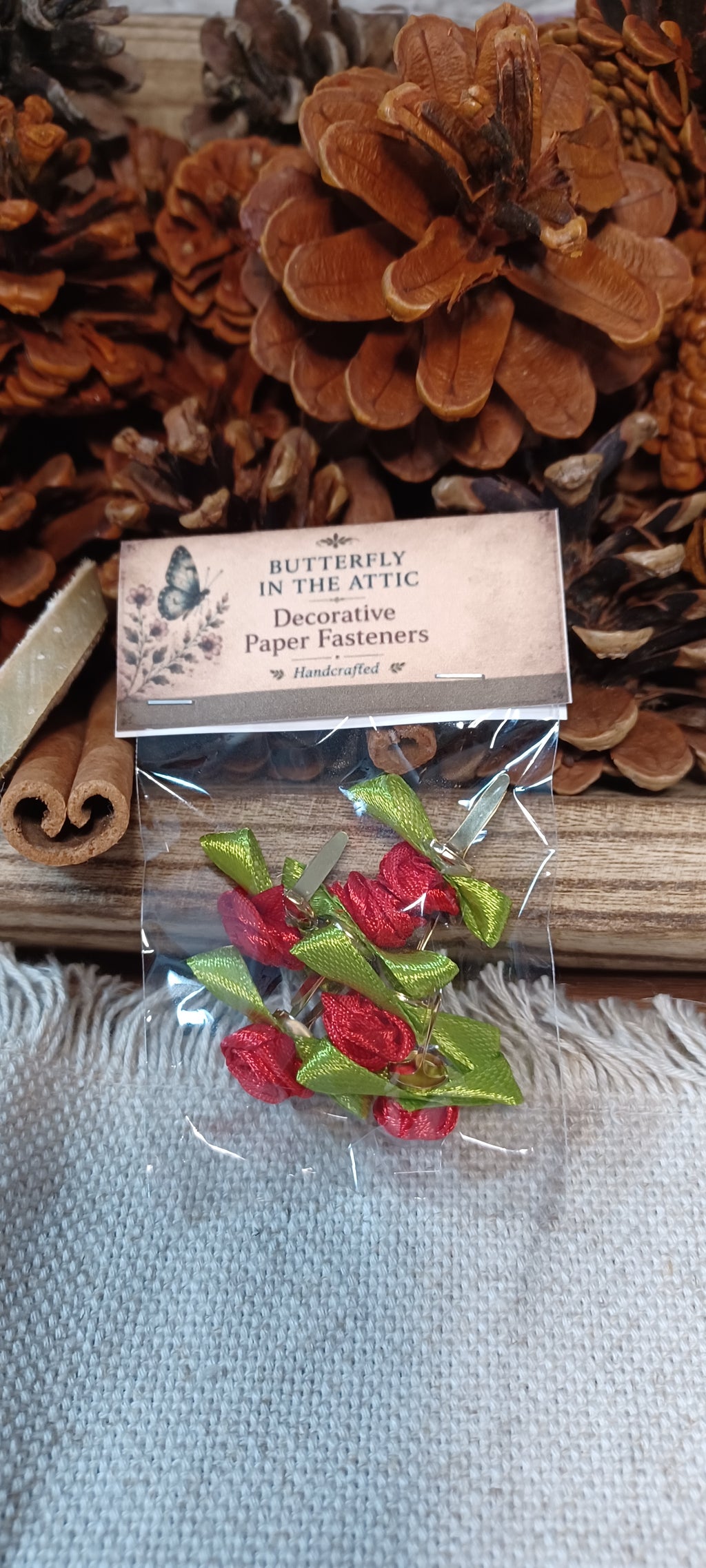 A small clear plastic bag containing red decorative paper fasteners with green ribbon stems, attached to a paper label that reads 'Butterfly in the Attic Decorative Paper Fasteners.' The bag is placed on a rustic wooden surface with pinecones and cinnamon sticks in the background.