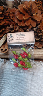 A small clear plastic bag containing red decorative paper fasteners with green ribbon stems, attached to a paper label that reads 'Butterfly in the Attic Decorative Paper Fasteners.' The bag is placed on a rustic wooden surface with pinecones and cinnamon sticks in the background.
