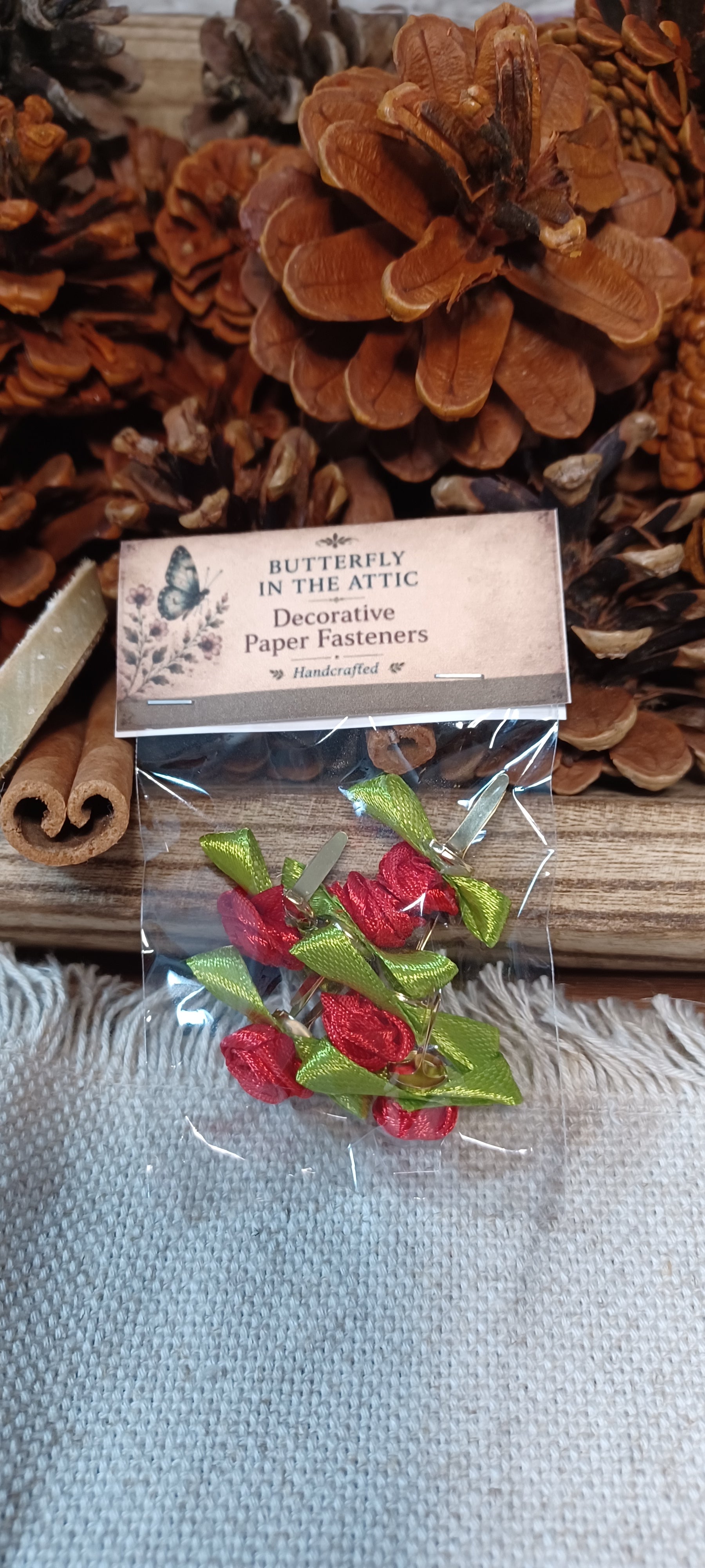A small clear plastic bag containing red decorative paper fasteners with green ribbon stems, attached to a paper label that reads 'Butterfly in the Attic Decorative Paper Fasteners.' The bag is placed on a rustic wooden surface with pinecones and cinnamon sticks in the background.