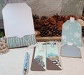A coordinated set of winter-themed paper tags, bookmarks, and a matching pen arranged on a textured fabric surface with pinecones in the background. The tags and bookmarks feature pale aqua, white snowflakes, brown geometric accents, and printed scenic artwork, while one tag is plain white with a snowflake pattern.