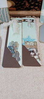 Two decorative paper bookmarks with brown triangular lower sections and illustrated top sections, placed on a beige fabric surface.