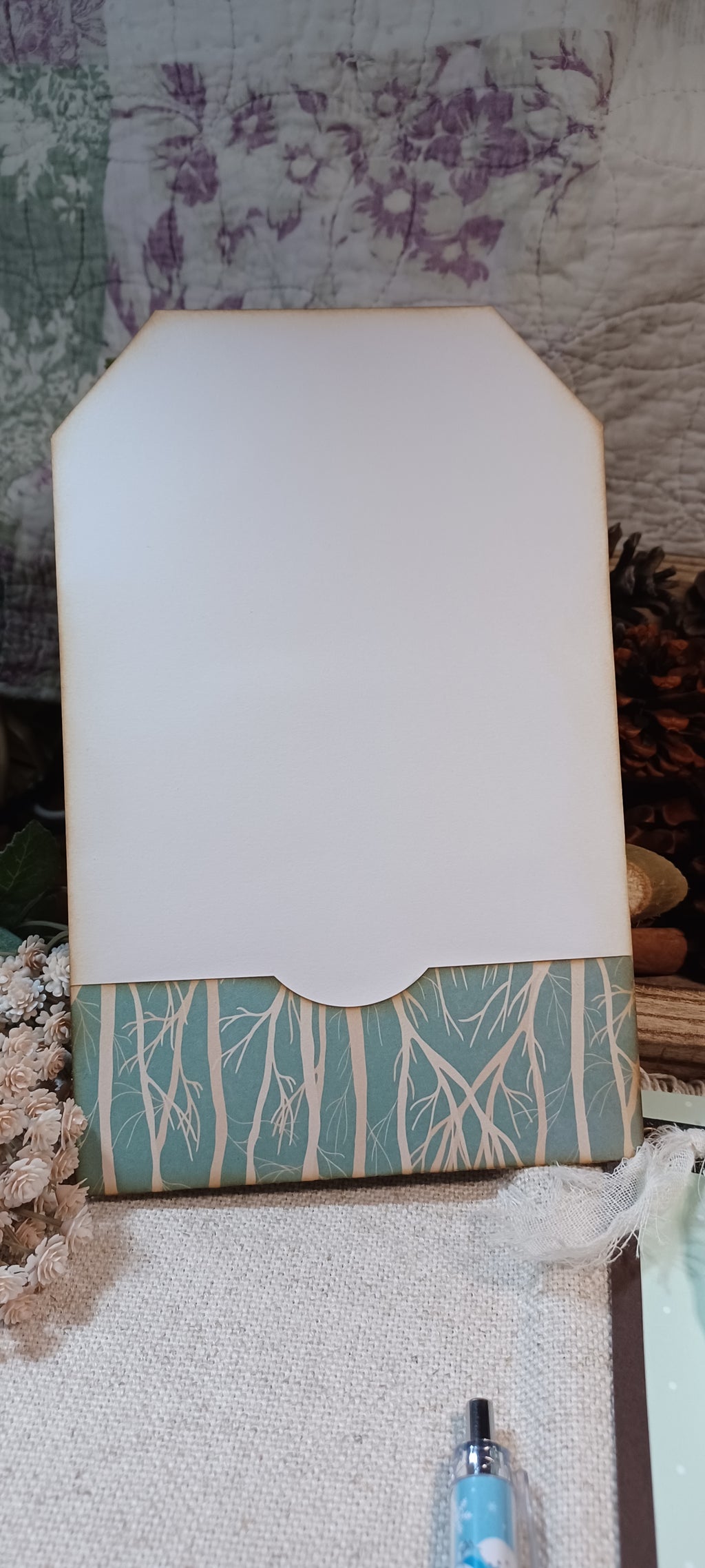 A handmade paper tag or card with a white top section and a patterned bottom border in teal and cream, styled on a textured fabric surface with dried flowers and a pen nearby.