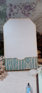 A handmade paper tag or card with a white top section and a patterned bottom border in teal and cream, styled on a textured fabric surface with dried flowers and a pen nearby.