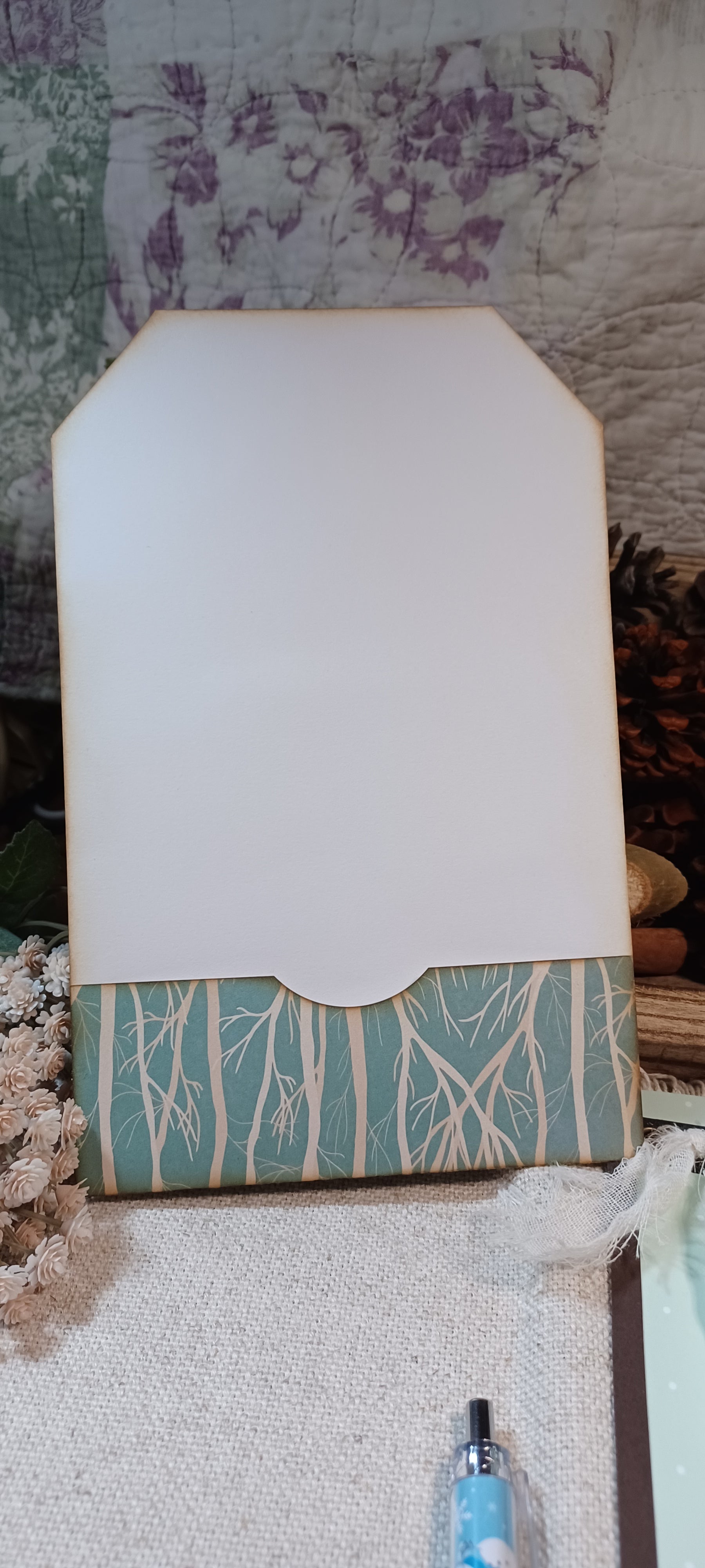 A handmade paper tag or card with a white top section and a patterned bottom border in teal and cream, styled on a textured fabric surface with dried flowers and a pen nearby.