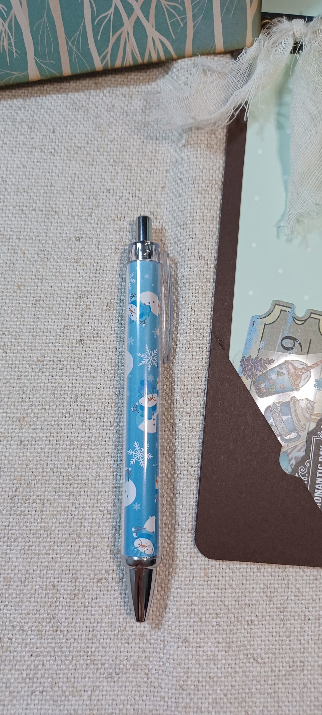 A light blue retractable pen with a winter-themed pattern of snowflakes, polar bears, and snow globes, resting on a textured beige fabric surface next to a brown card.