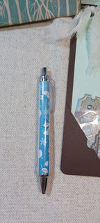 A light blue retractable pen with a winter-themed pattern of snowflakes, polar bears, and snow globes, resting on a textured beige fabric surface next to a brown card.