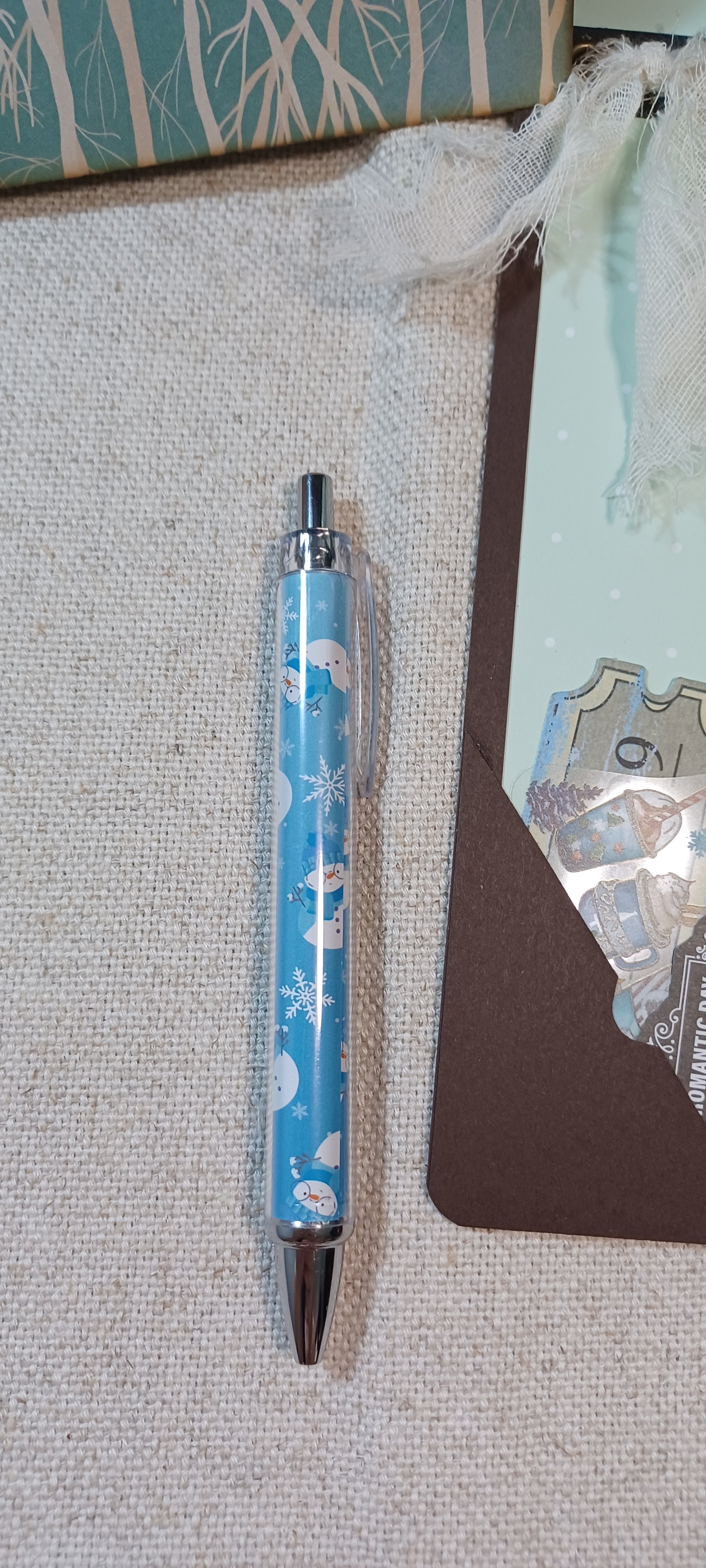 A light blue retractable pen with a winter-themed pattern of snowflakes, polar bears, and snow globes, resting on a textured beige fabric surface next to a brown card.