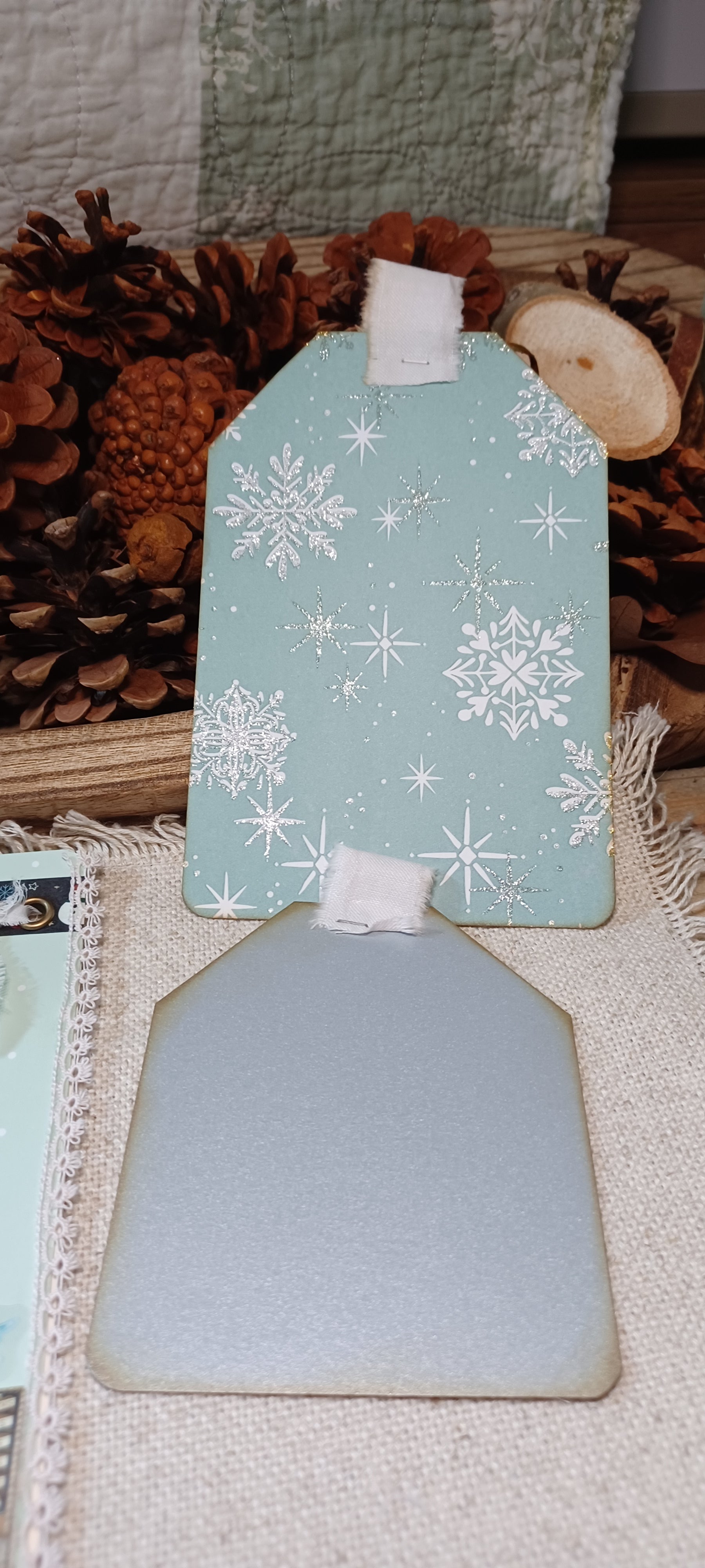 Two decorative gift tags with a winter theme, one in light blue with white snowflakes and the other in a matte sage green, displayed on a burlap surface with pinecones in the background.