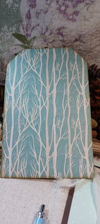 A rectangular decorative sign or plaque with an arched top, featuring a light blue background with a repeating pattern of white leafless tree branches. The sign is photographed on a styled surface with a quilted blanket, burlap fabric, and a small plant in the background.