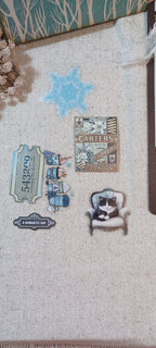 A collection of vintage-style enamel pins displayed on a textured beige fabric surface. The pins include designs such as a blue snowflake, a 'CARTERS' luggage tag, a ticket reading '54329', a small blue and white vintage train set, a black-and-white cat in an armchair, and a 'A ROMANTIC DAY' label.