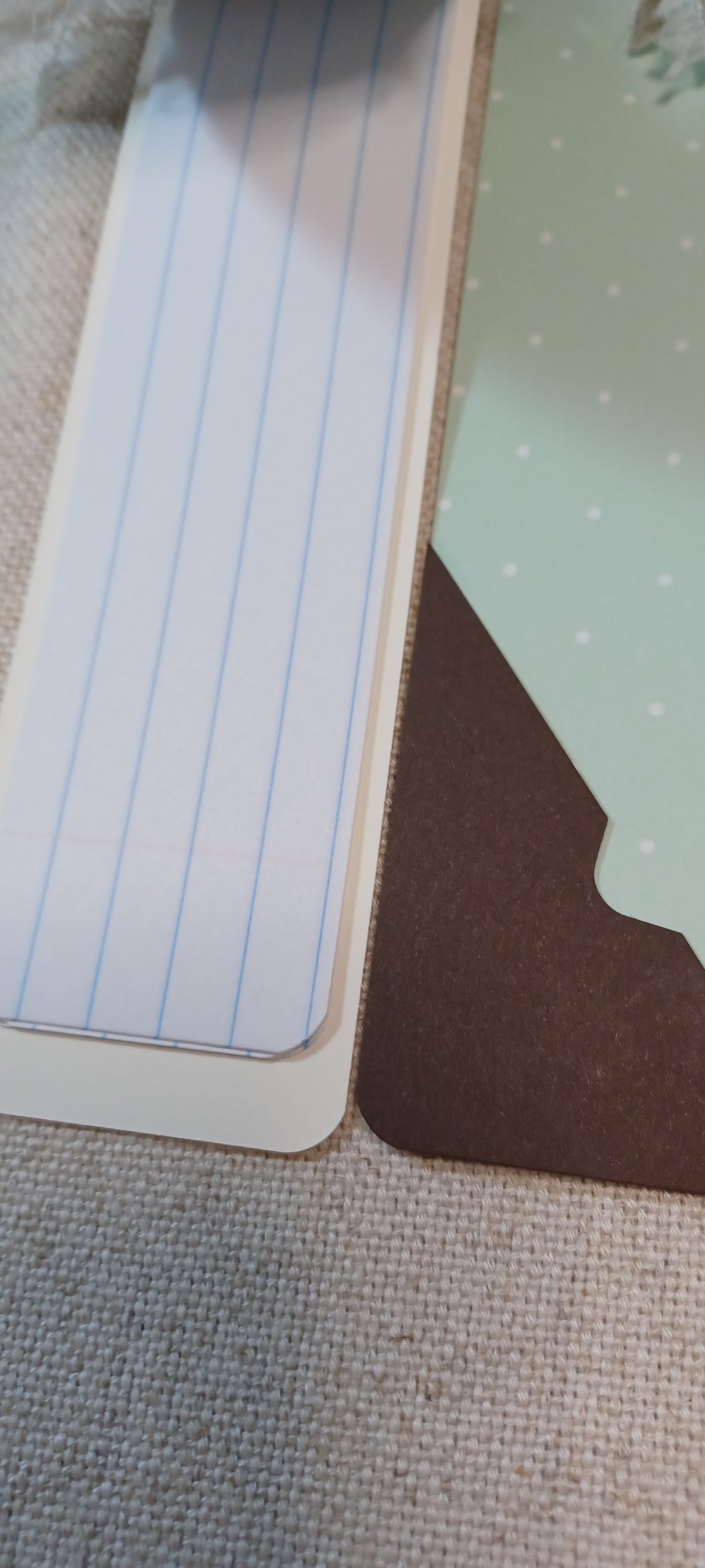 A close-up of a white lined notepad page with blue vertical lines, placed next to a brown paper tab and a mint green patterned sheet on a textured beige fabric surface.