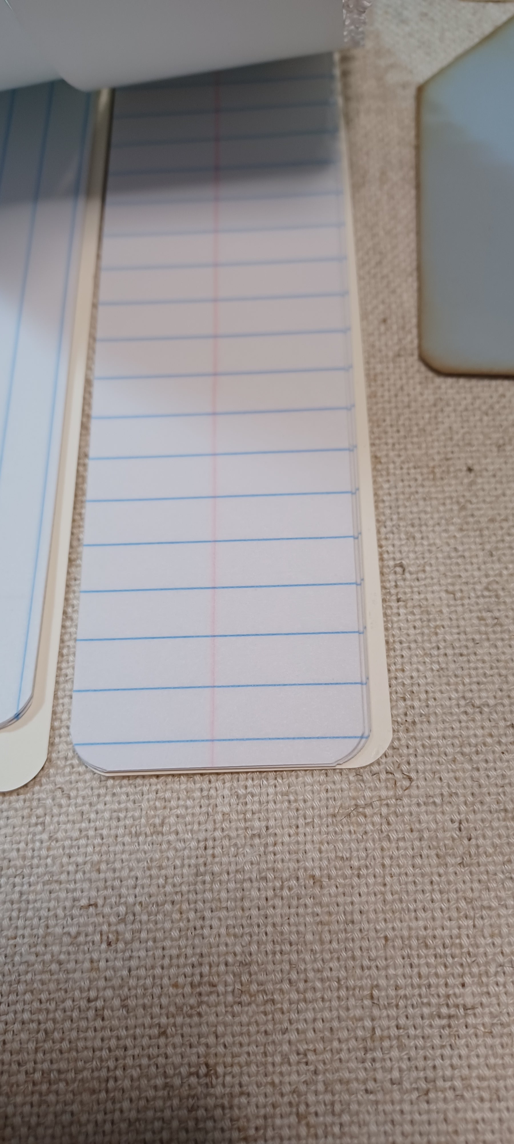 A close-up of lined paper notepad pages with blue and red lines, partially visible on a beige woven fabric surface.