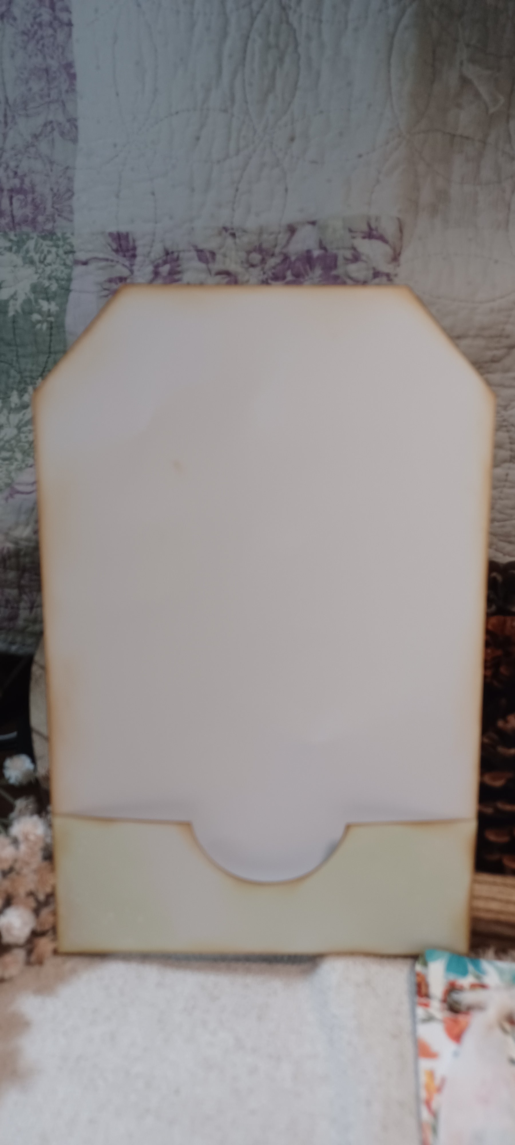 A handmade wooden card holder or envelope with a scalloped cream-colored front flap and a light wood border, set against a patchwork quilt background.