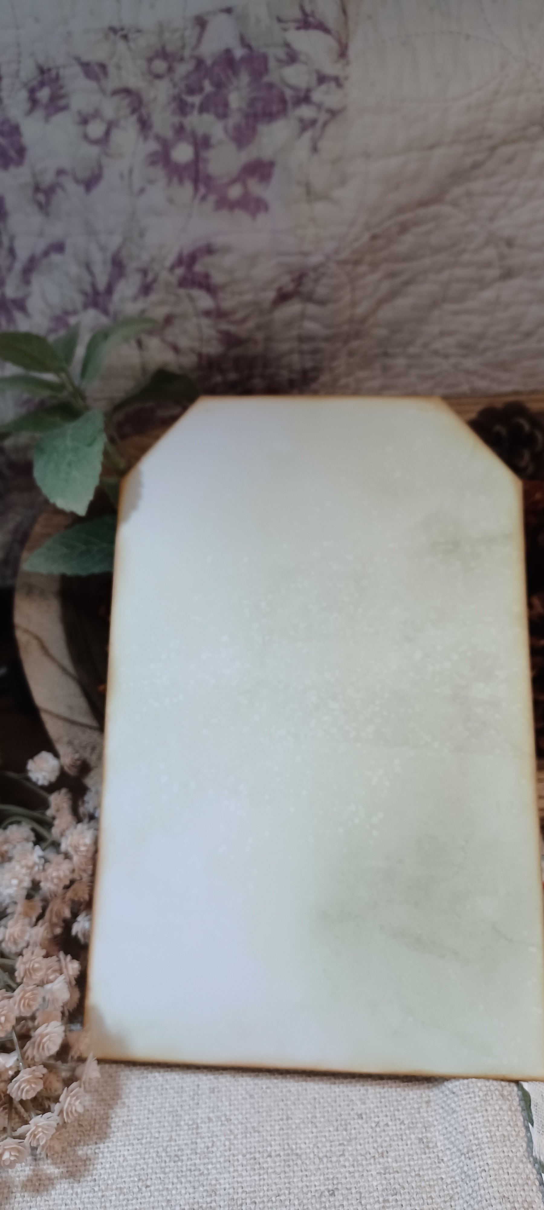 A blank, off-white rectangular card or sign with rounded corners, displayed on a textured fabric surface next to dried flowers and a floral-patterned background.