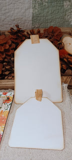 Two blank white wooden tag-shaped photo frames with natural wood edges and small tan tabs at the top, displayed on a burlap surface with autumn decor in the background.